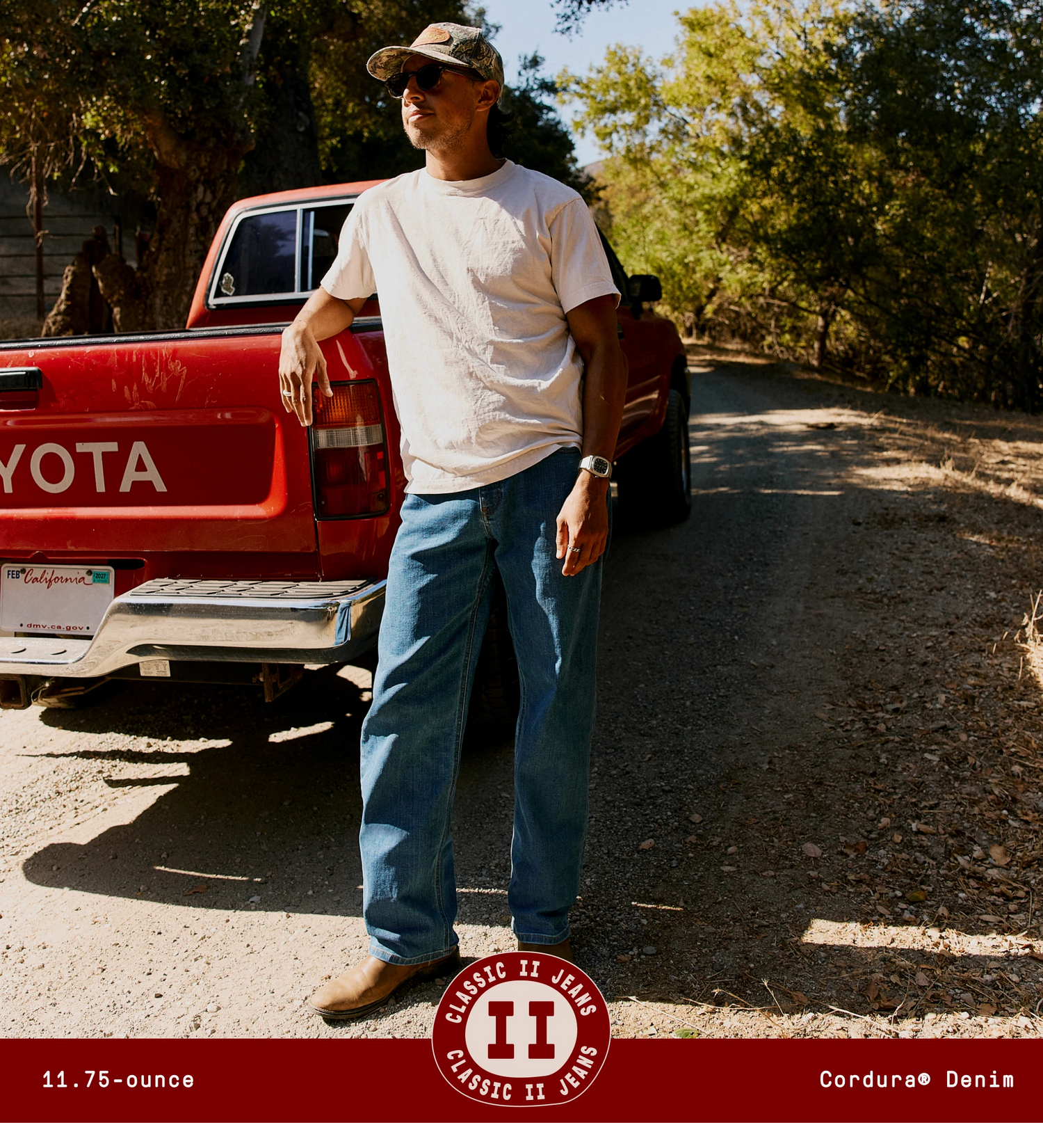 A man wearing a white t-shirt, blue jeans, and brown boots stands on a dirt road next to a red Toyota pickup truck. A circular logo for 'Classic II Jeans' is displayed at the bottom, along with the text "11.75-ounce" and "Cordura Denim"