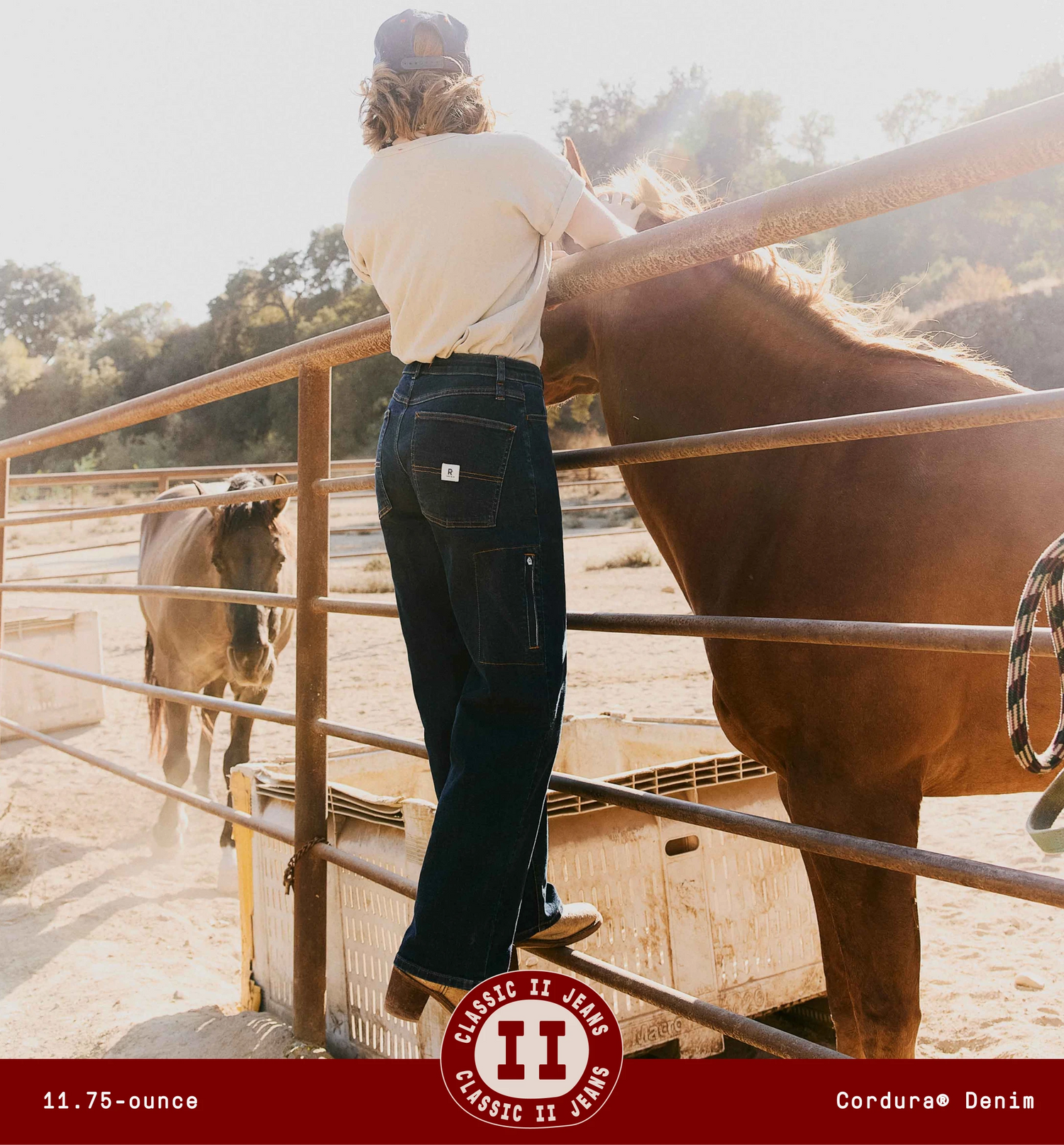 Woman wearing dark-wash denim jeans standing on the fence at a sunlit horse corral, shown from the back.