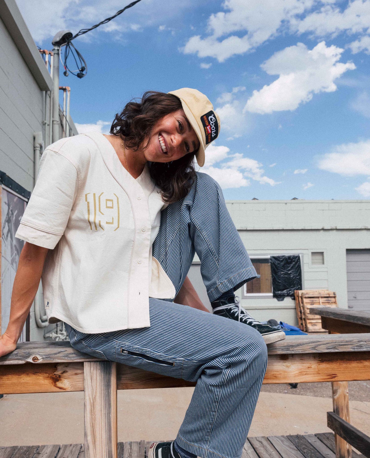 A woman sitting on a wooden bench outdoors under a blue sky, wearing a cream baseball-style shirt with gold lettering, a blue striped button-up shirt, blue striped pants, and a beige cap with a black logo patch.