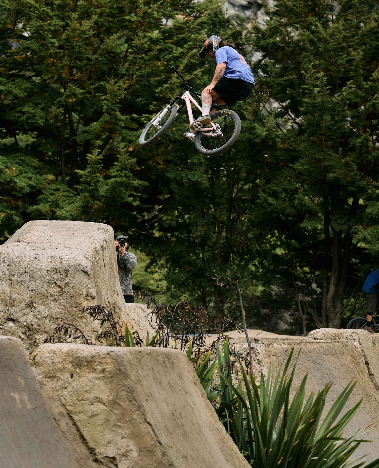 Person on a bicycle performing a jump in an outdoor setting with trees and concrete structures.
