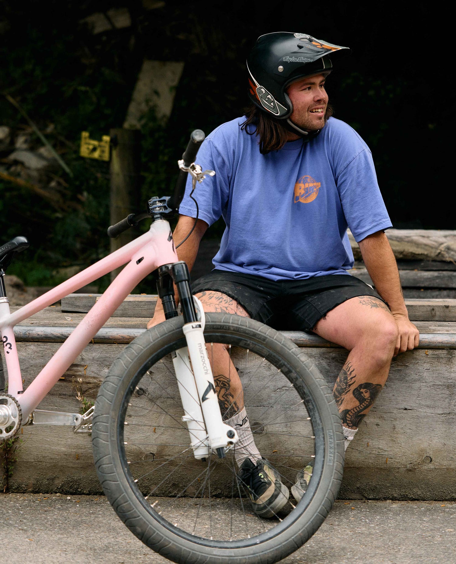 Person sitting on a wooden log with a pink bicycle, wearing a helmet and blue shirt.