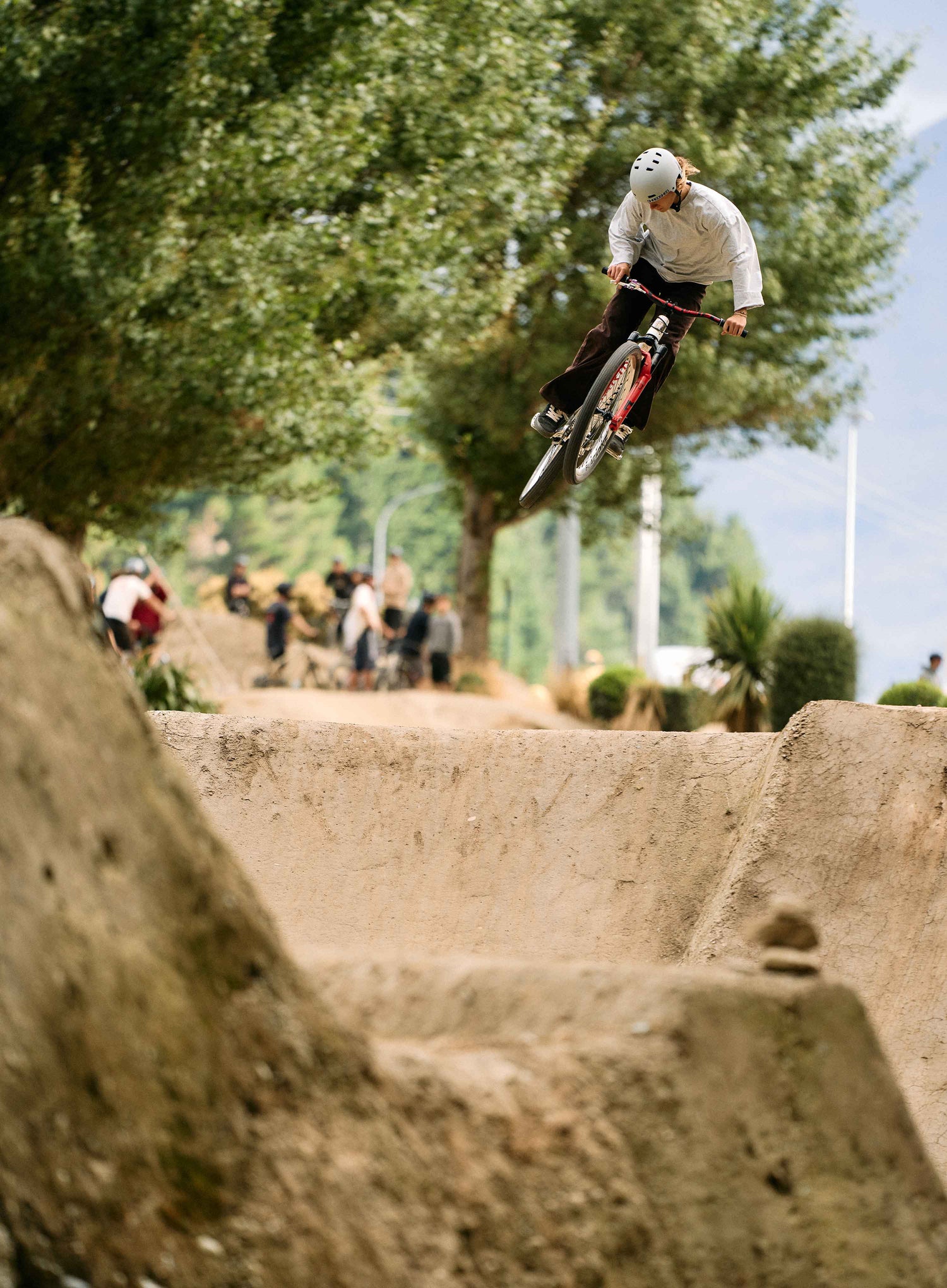 Person on a bicycle performing a jump in an outdoor setting with trees and people in the background.