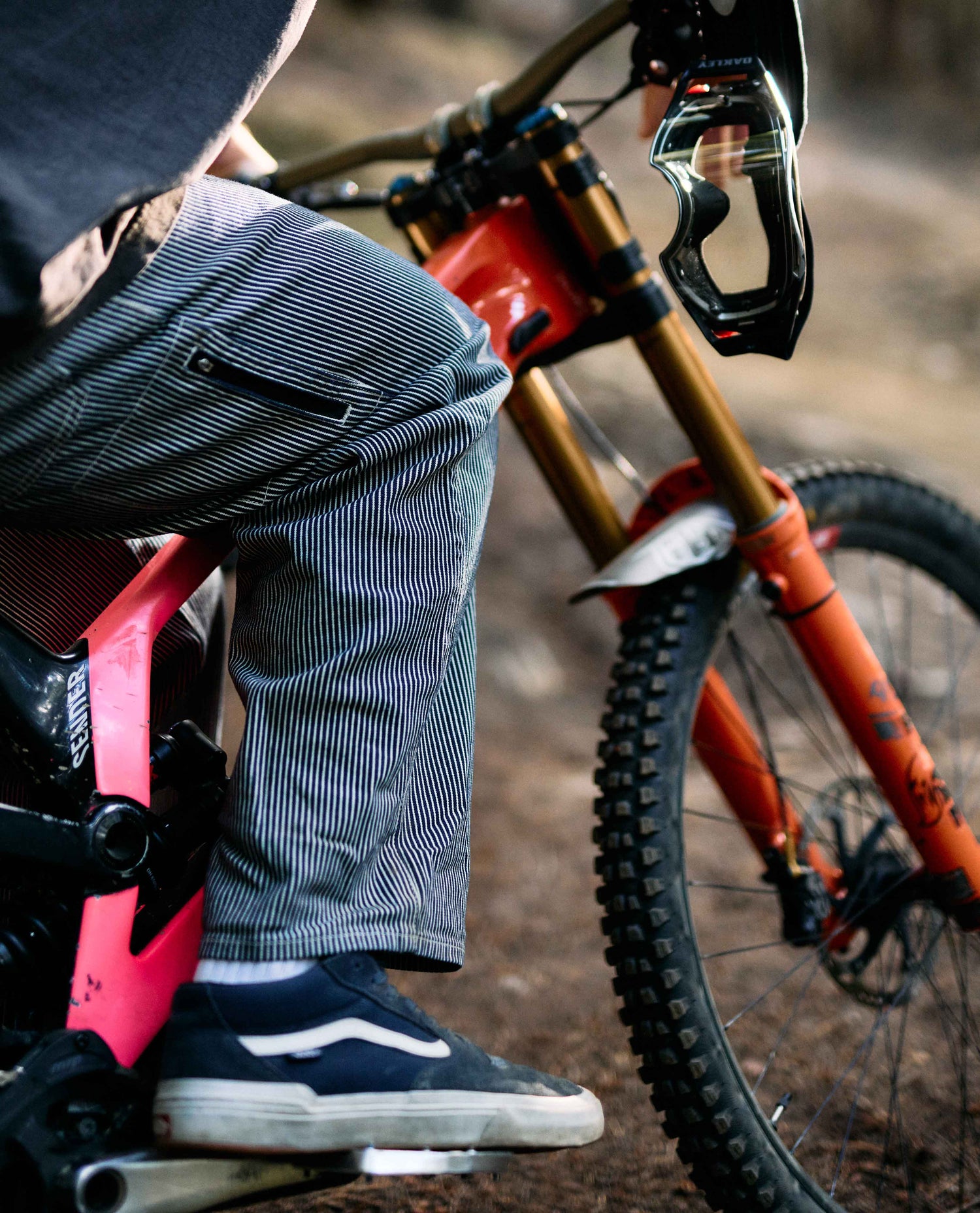 Person wearing striped pants and black shoes next to a red bicycle on a dirt path