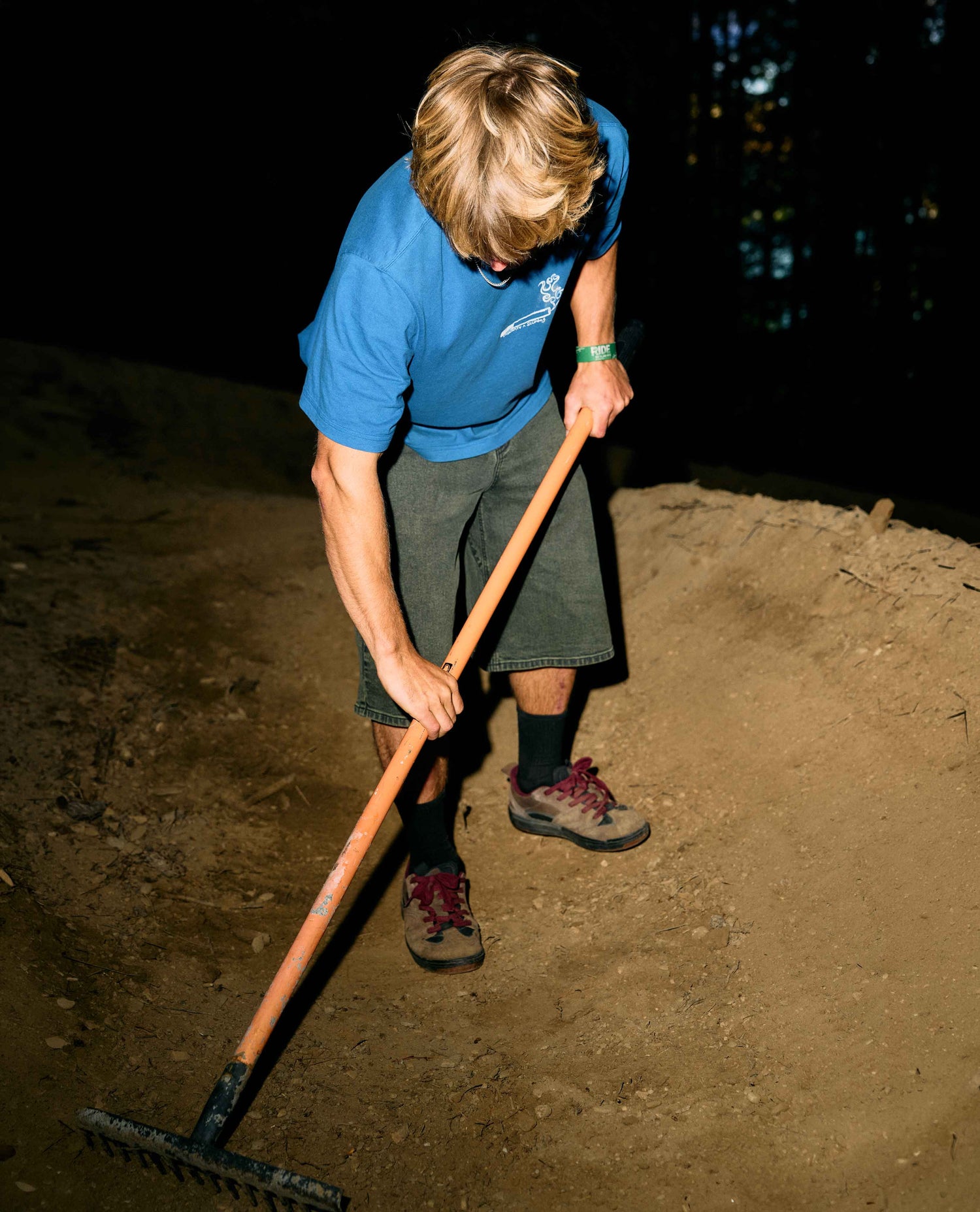 Person in blue shirt and shorts using a rake on a dirt surface at night.