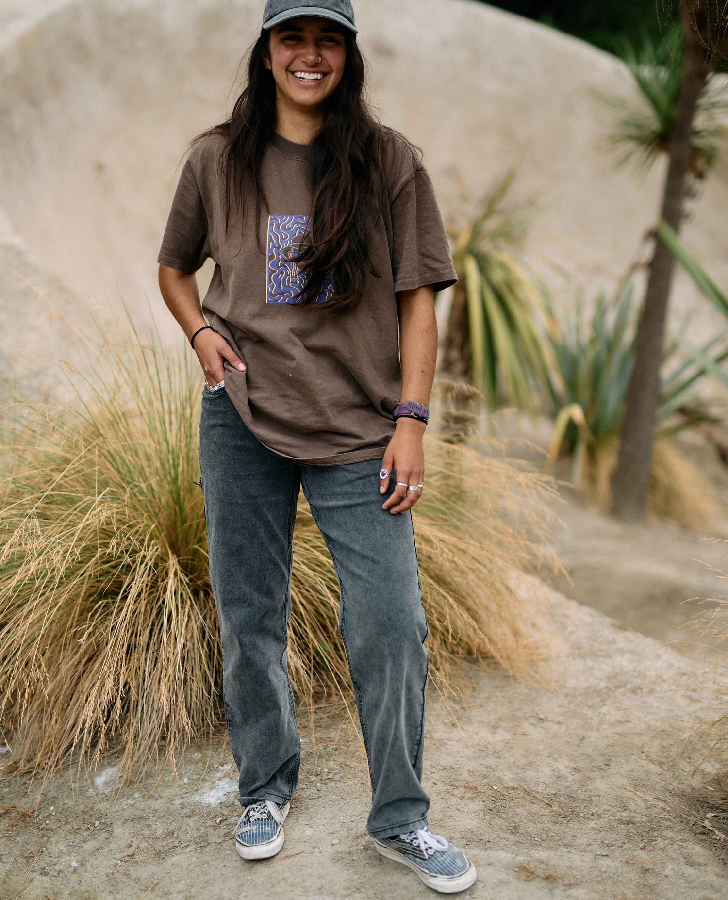 Person wearing a brown t-shirt with a graphic design, blue jeans, and a cap, standing in a natural setting.