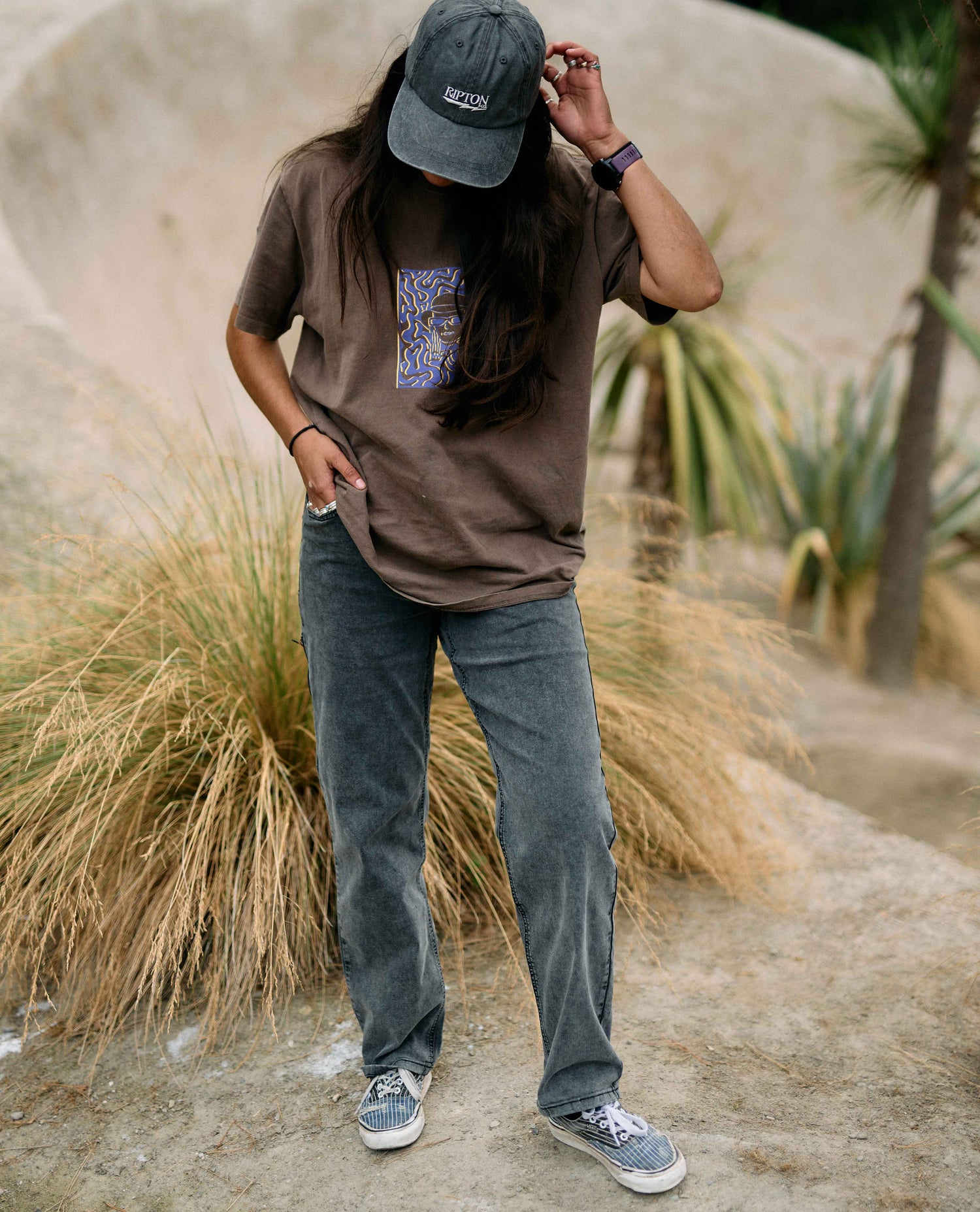 Person wearing a brown t-shirt with a graphic design, blue jeans, and a cap in a natural setting.