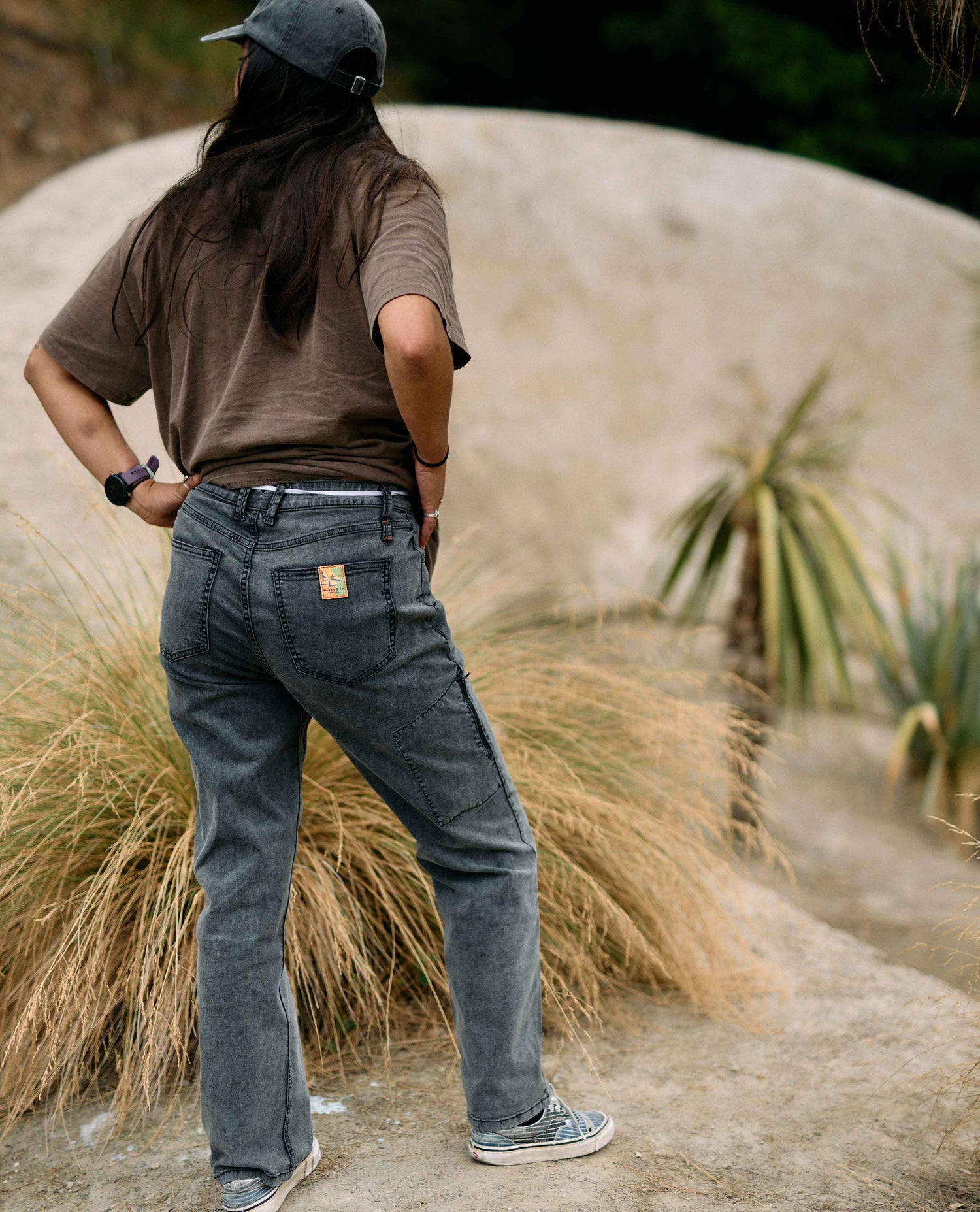 Person wearing a brown shirt and gray pants standing on a path with plants and sand in the background