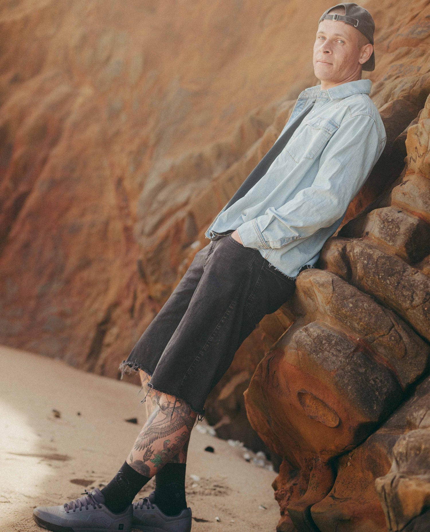 Man in a light denim shirt and black shorts leaning against a rock with a beachy landscape in the background