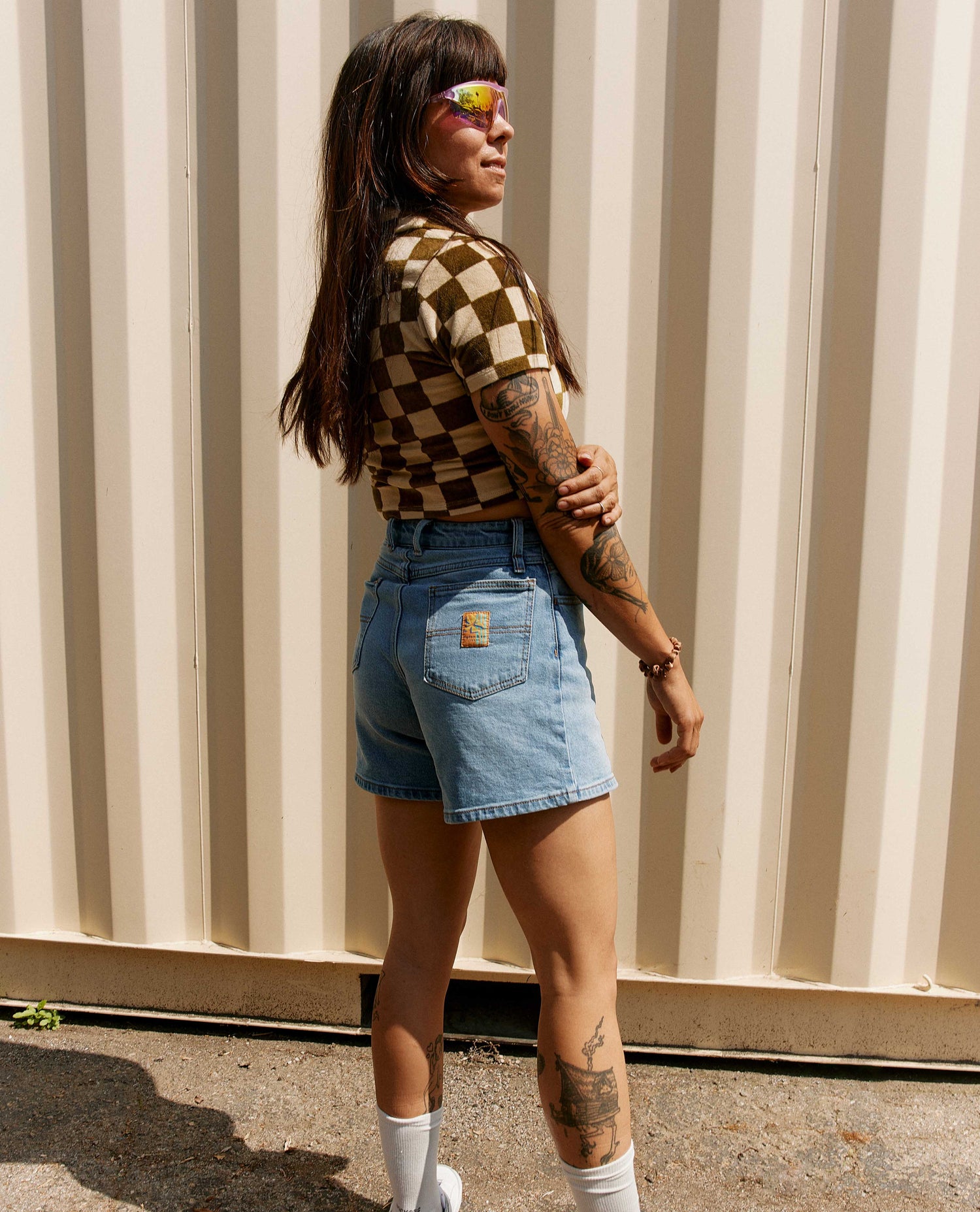 Woman standing in front of a shipping crate wearing a checkered crop top and jean shorts with white shoes. 