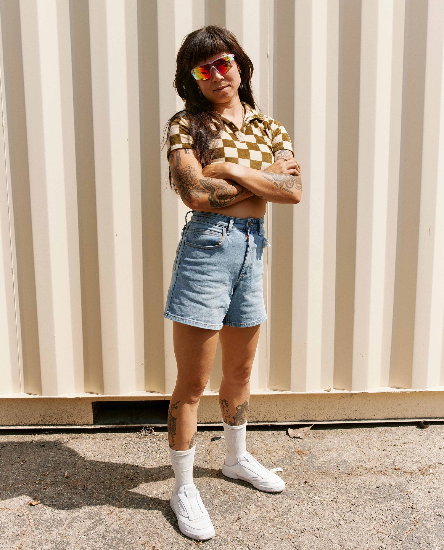 Woman standing in front of a shipping crate wearing a checkered crop top and jean shorts with white shoes. 